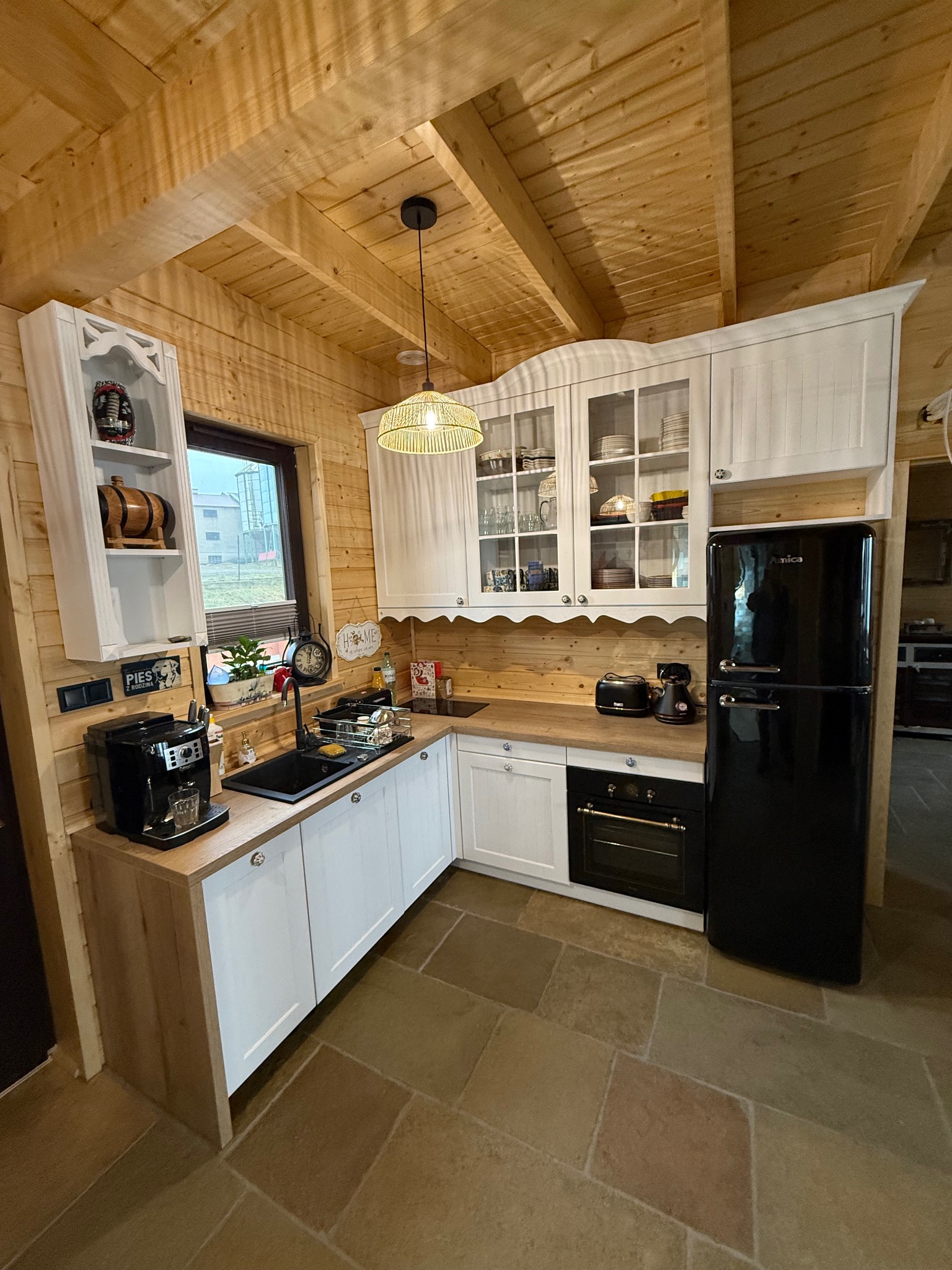 Drewniana kuchnia z białymi szafkami, czarnym sprzętem AGD i kamienną podłogą, oświetlona wiklinową lampą.