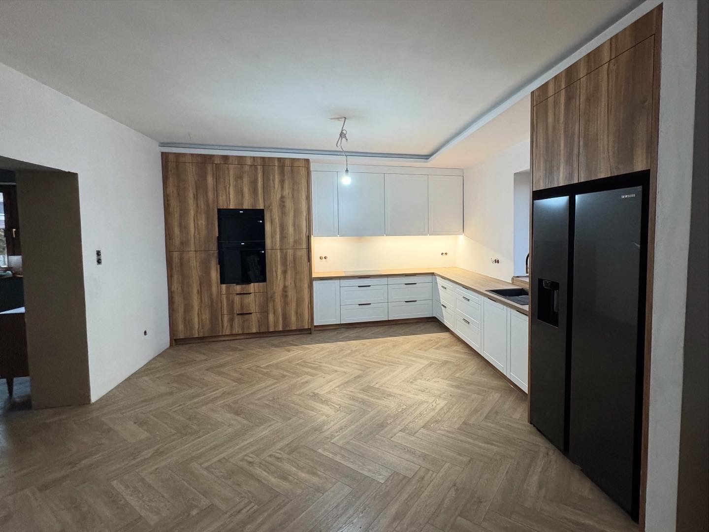 Jasna kuchnia w stylu skandynawskim z drewnianymi blatami i parkietem w jodełkę, zabudowa z piekarnikiem i lodówką side-by-side, oświetlenie podszafkowe.