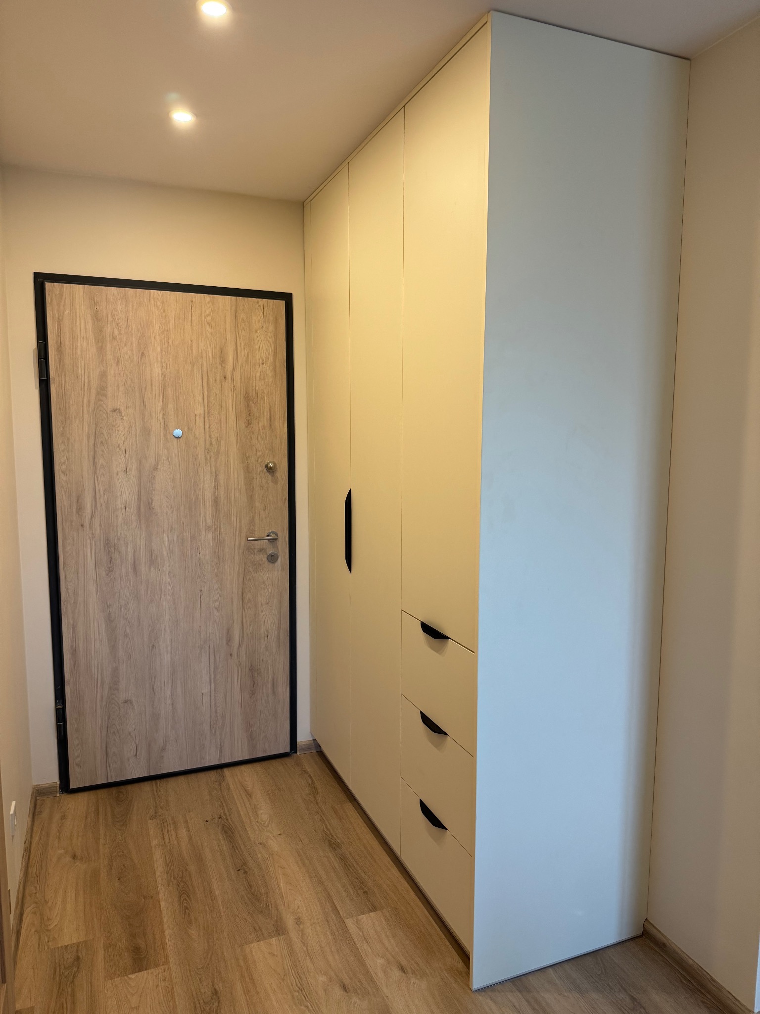 Jasna szafa z szufladami i długim uchwytem obok drzwi wejściowych z drewnopodobnym wykończeniem i czarną ramą, widok na korytarz z podłogą imitującą drewno.
