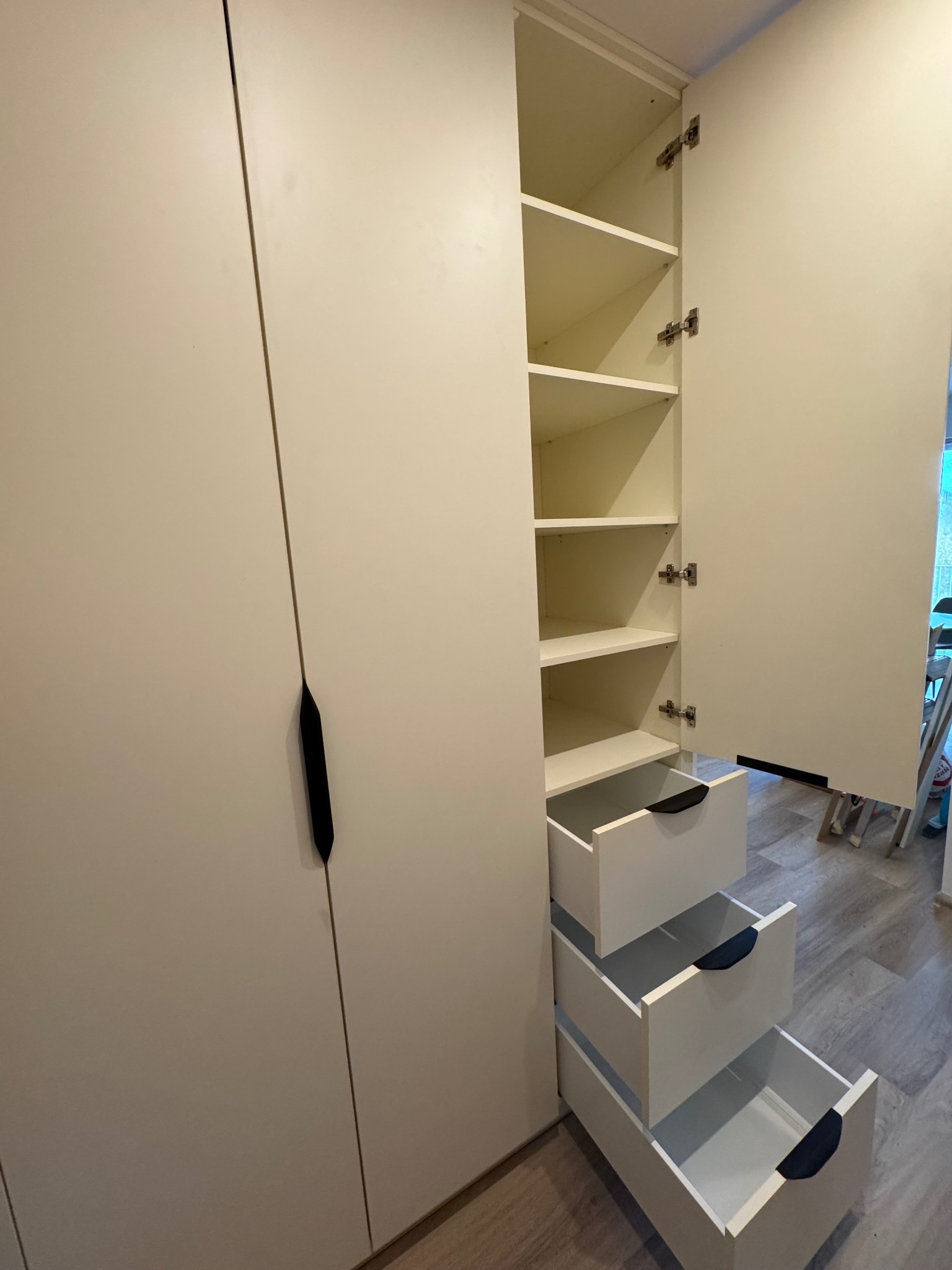 Biała szafa z otwartymi drzwiami i trzema wysuniętymi szufladami z czarnymi uchwytami, widoczne półki wewnątrz.