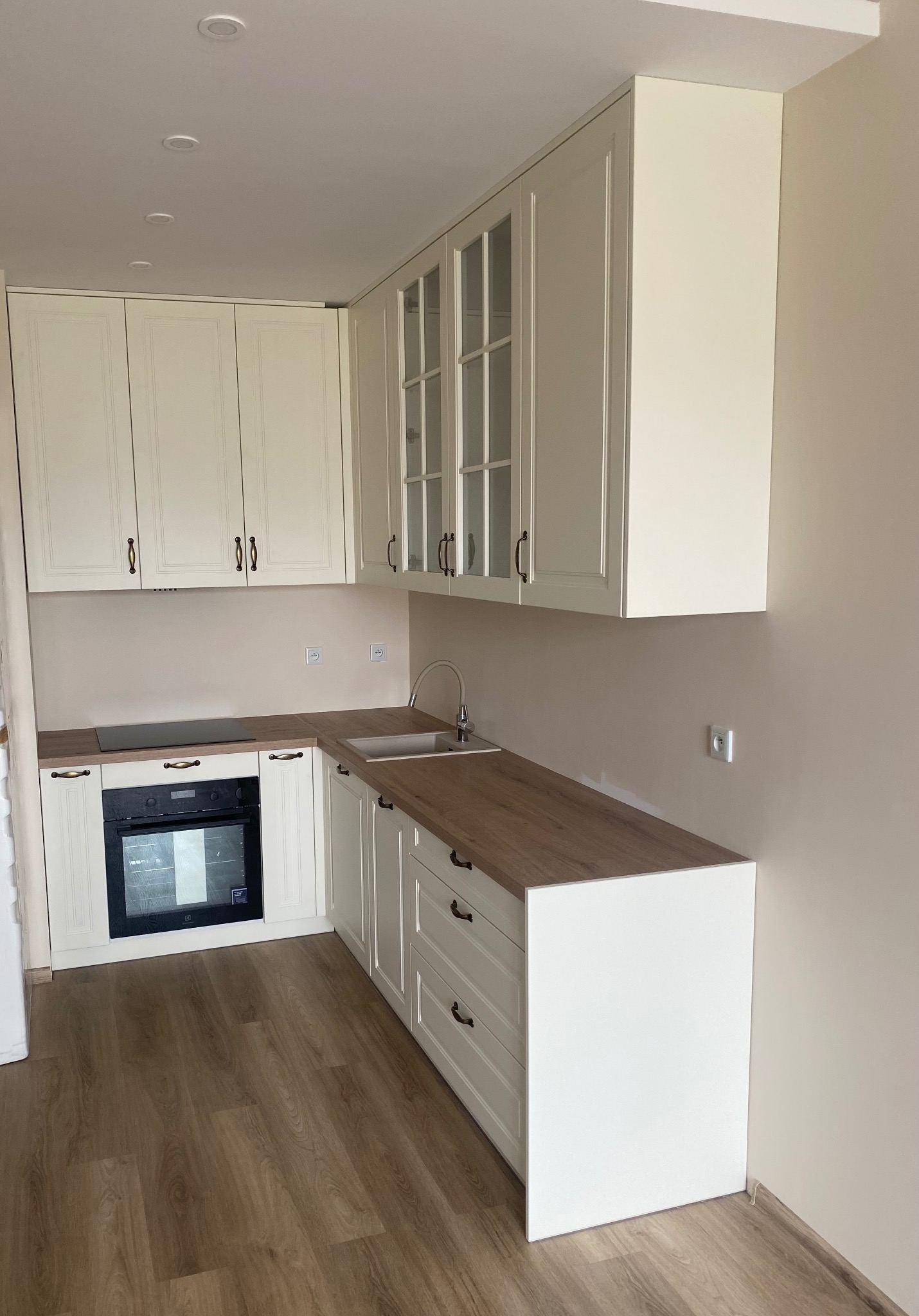 Jasna kuchnia z drewnianym blatem, fronty szafek frezowane, widoczne szklane witryny w górnych szafkach, brązowe uchwyty, płyta indukcyjna i piekarnik w zabudowie.