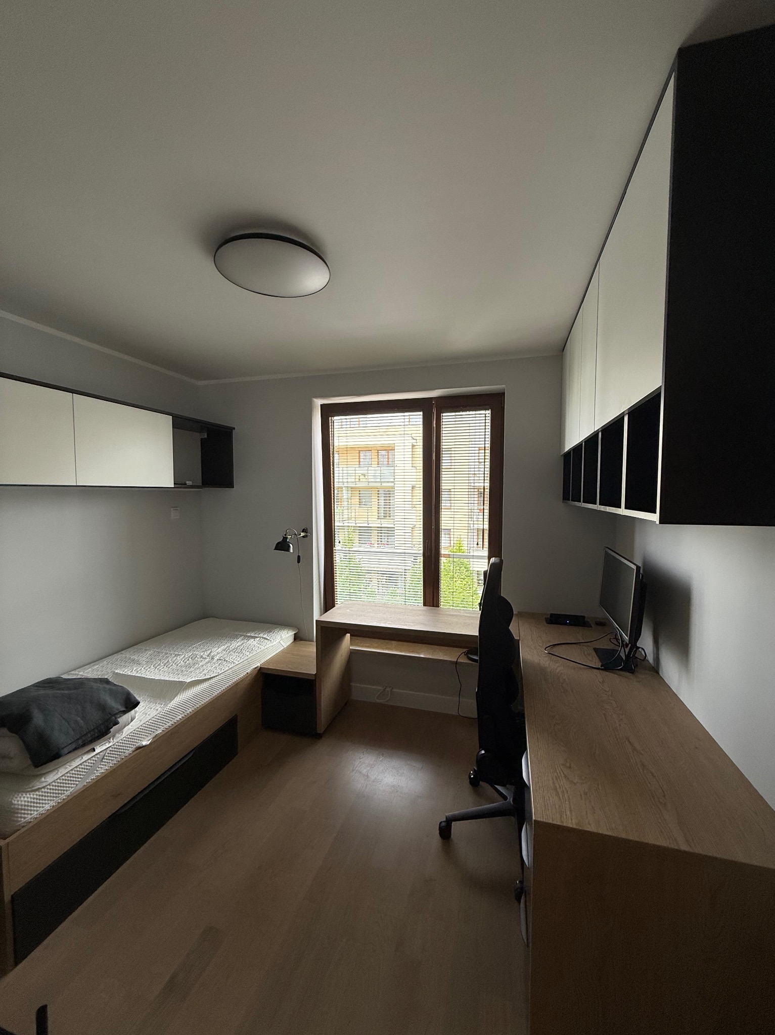 Pokój z zabudową meblową: biurko pod oknem, szafki wiszące nad łóżkiem i długi blat z szafkami na ścianie. Meble w odcieniach bieli i drewna.