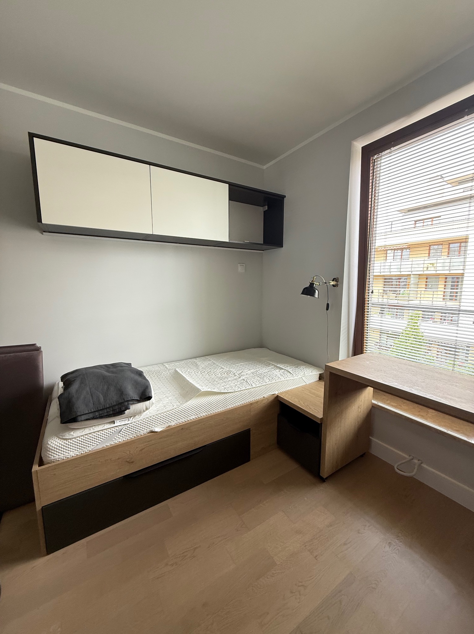 Pokój z drewnianym łóżkiem z szufladami, biurkiem przy oknie i szafkami wiszącymi w odcieniach bieli i czerni. Widok na budynek przez żaluzje.