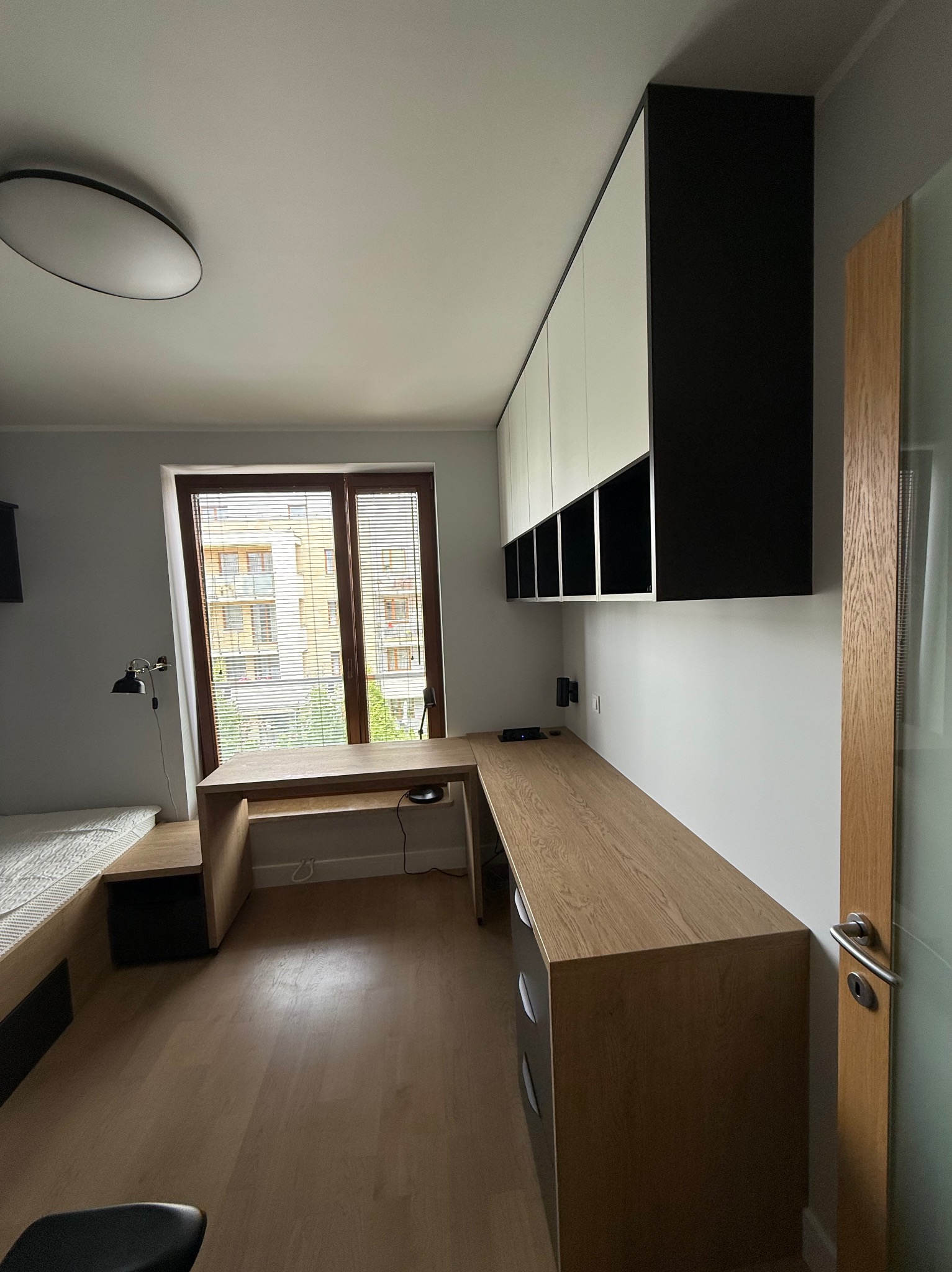 Pokój z biurkiem na wymiar z blatem dębowym, wiszącymi szafkami biało-czarnymi i łóżkiem z drewnianą ramą. Widok na budynek przez okno z żaluzjami.