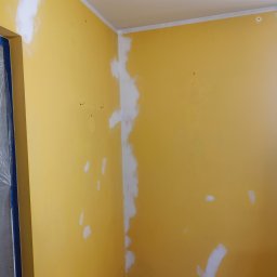 SEBRATECH Sebastian Raczyński - Żółta ściana w trakcie przygotowania do malowania, z widocznymi miejscami po łataniu i szpachlowaniu, zabezpieczone folią drzwi.