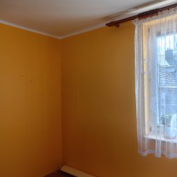 SEBRATECH Sebastian Raczyński - Pokój z pomarańczowymi ścianami, drewnianą podłogą i oknem z firanką, widoczne ślady po poprzednich dekoracjach na ścianach, listwa przypodłogowa częściowo odklejona.