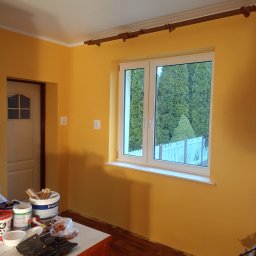 SEBRATECH Sebastian Raczyński - Pokój pomalowany na żółto, widoczne narzędzia malarskie i puszki z farbą na stole, okno z widokiem na zielone drzewa.