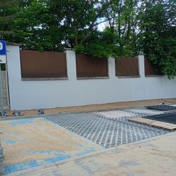 SEBRATECH Sebastian Raczyński - Układanie ażurowych płyt na parkingu. Widoczny piasek, palety i narzędzia przy białym murze z brązowymi elementami. Prace w toku, teren zielony w tle.