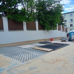 SEBRATECH Sebastian Raczyński - Układanie ażurowej kostki brukowej na posesji. Widoczne przygotowane elementy, palety i piasek. W tle ogrodzenie i budynek mieszkalny.