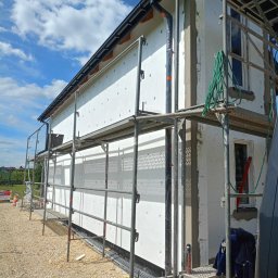 SEBRATECH Sebastian Raczyński - Dom w trakcie ocieplania: białe płyty izolacyjne na ścianach, rusztowanie, widoczny fragment elewacji z orynnowaniem. Słoneczny dzień, błękitne niebo.