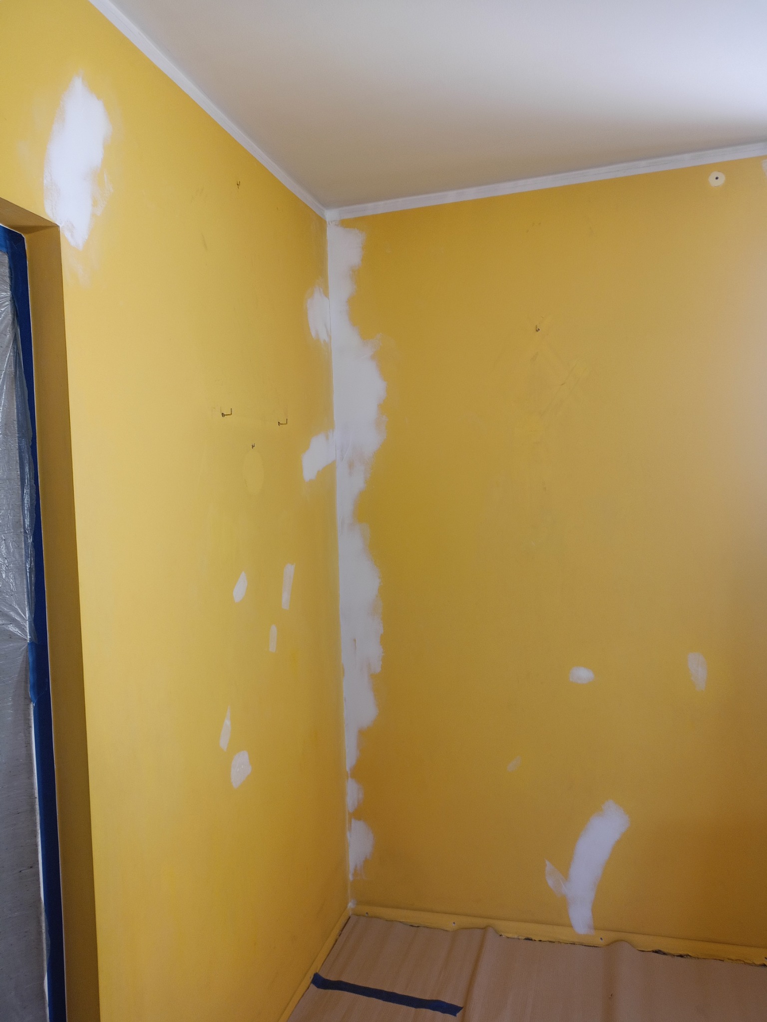 Żółta ściana w trakcie przygotowania do malowania, z widocznymi miejscami po łataniu i szpachlowaniu, zabezpieczone folią drzwi.