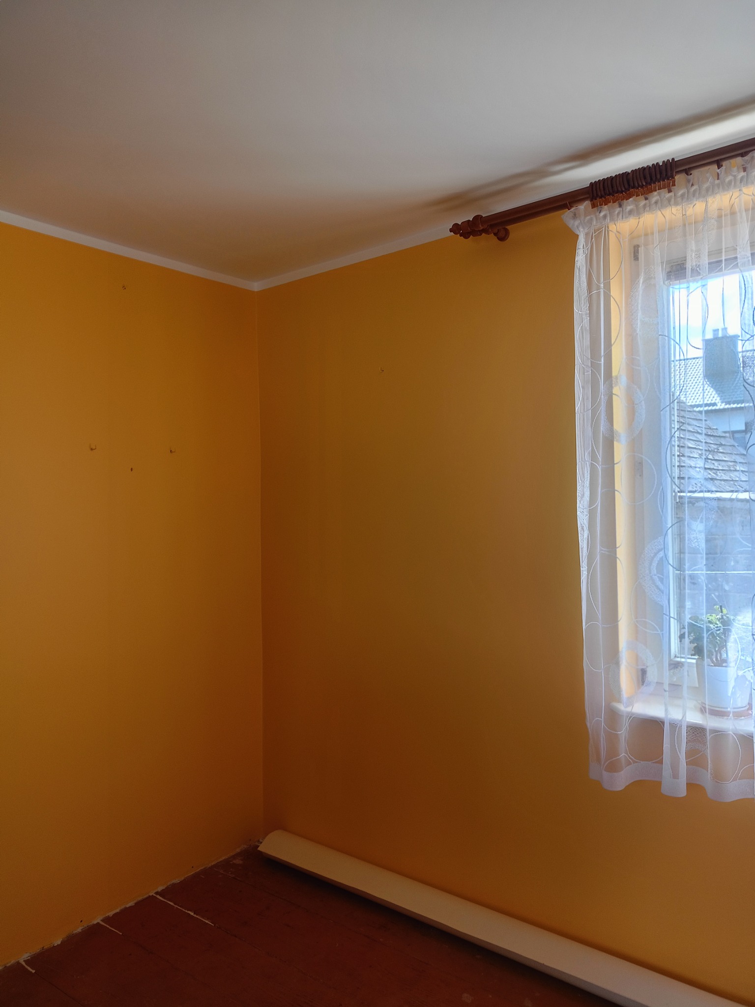 Pokój z pomarańczowymi ścianami, drewnianą podłogą i oknem z firanką, widoczne ślady po poprzednich dekoracjach na ścianach, listwa przypodłogowa częściowo odklejona.
