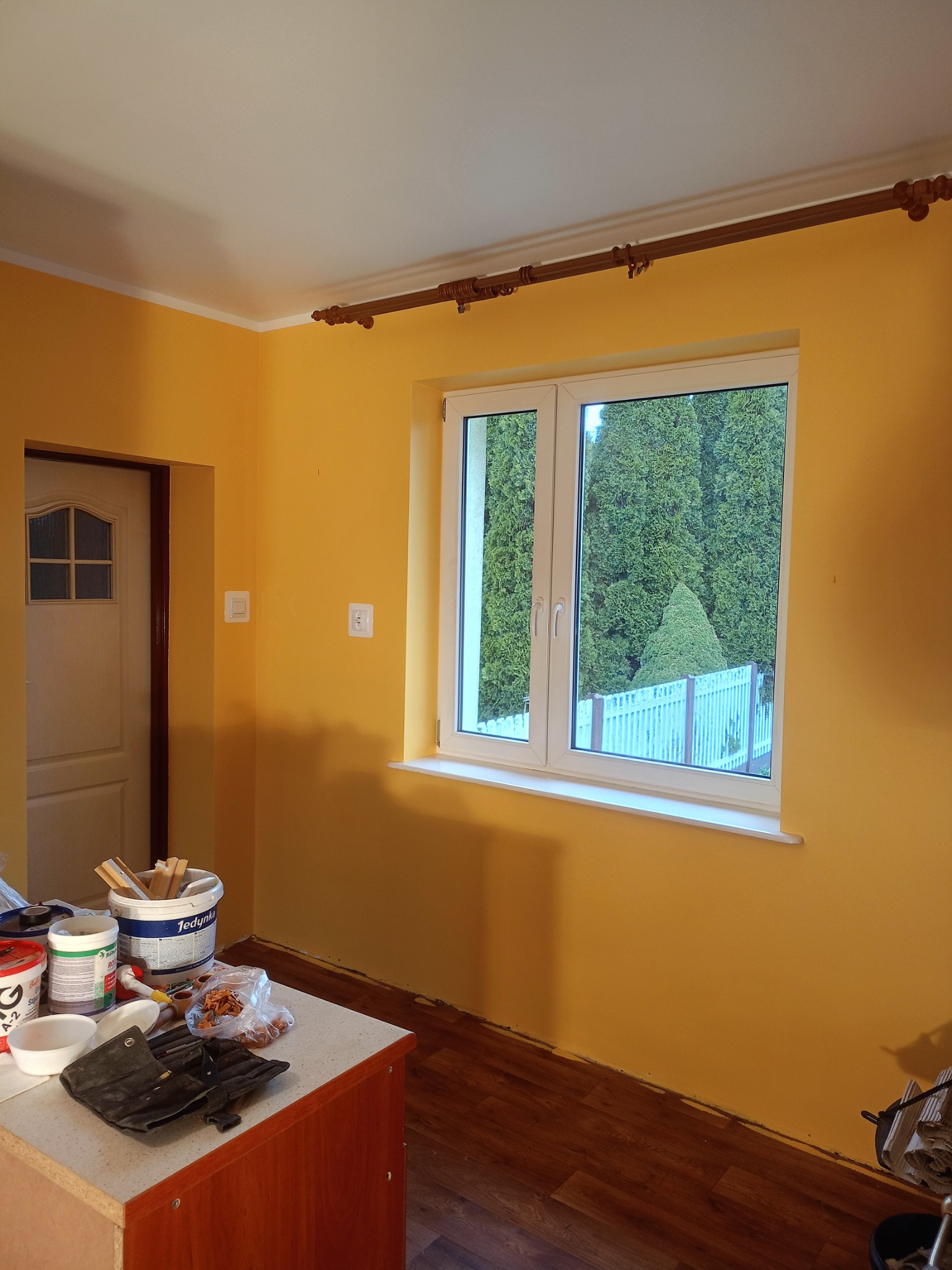 Pokój pomalowany na żółto, widoczne narzędzia malarskie i puszki z farbą na stole, okno z widokiem na zielone drzewa.