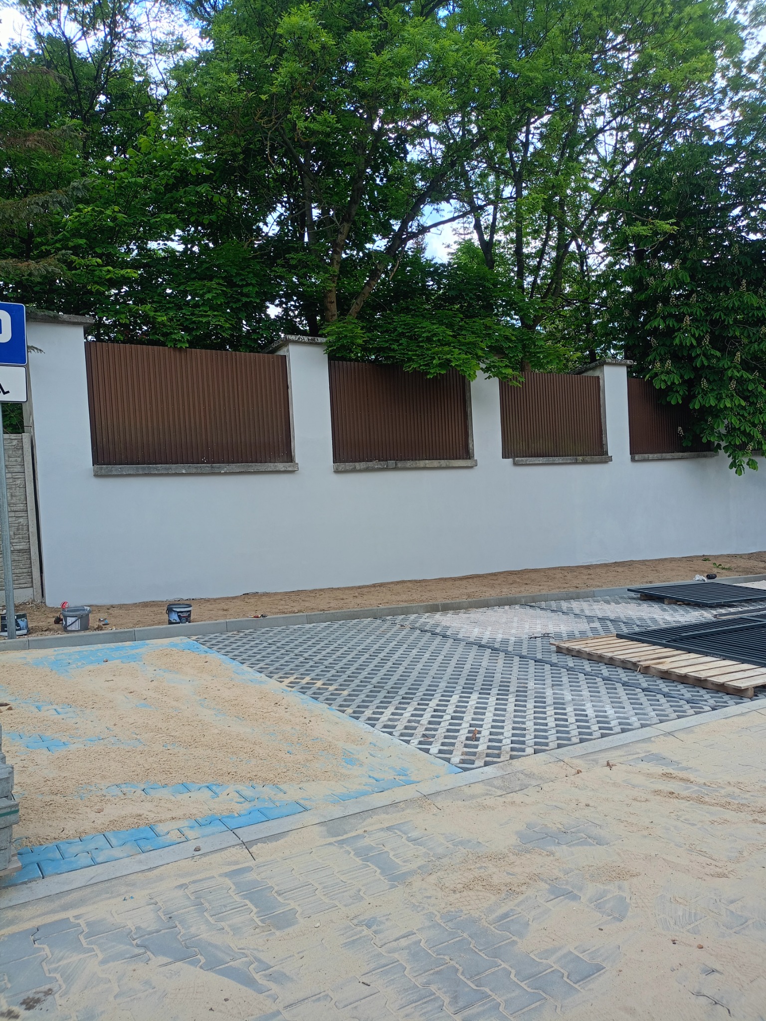 Układanie ażurowych płyt na parkingu. Widoczny piasek, palety i narzędzia przy białym murze z brązowymi elementami. Prace w toku, teren zielony w tle.