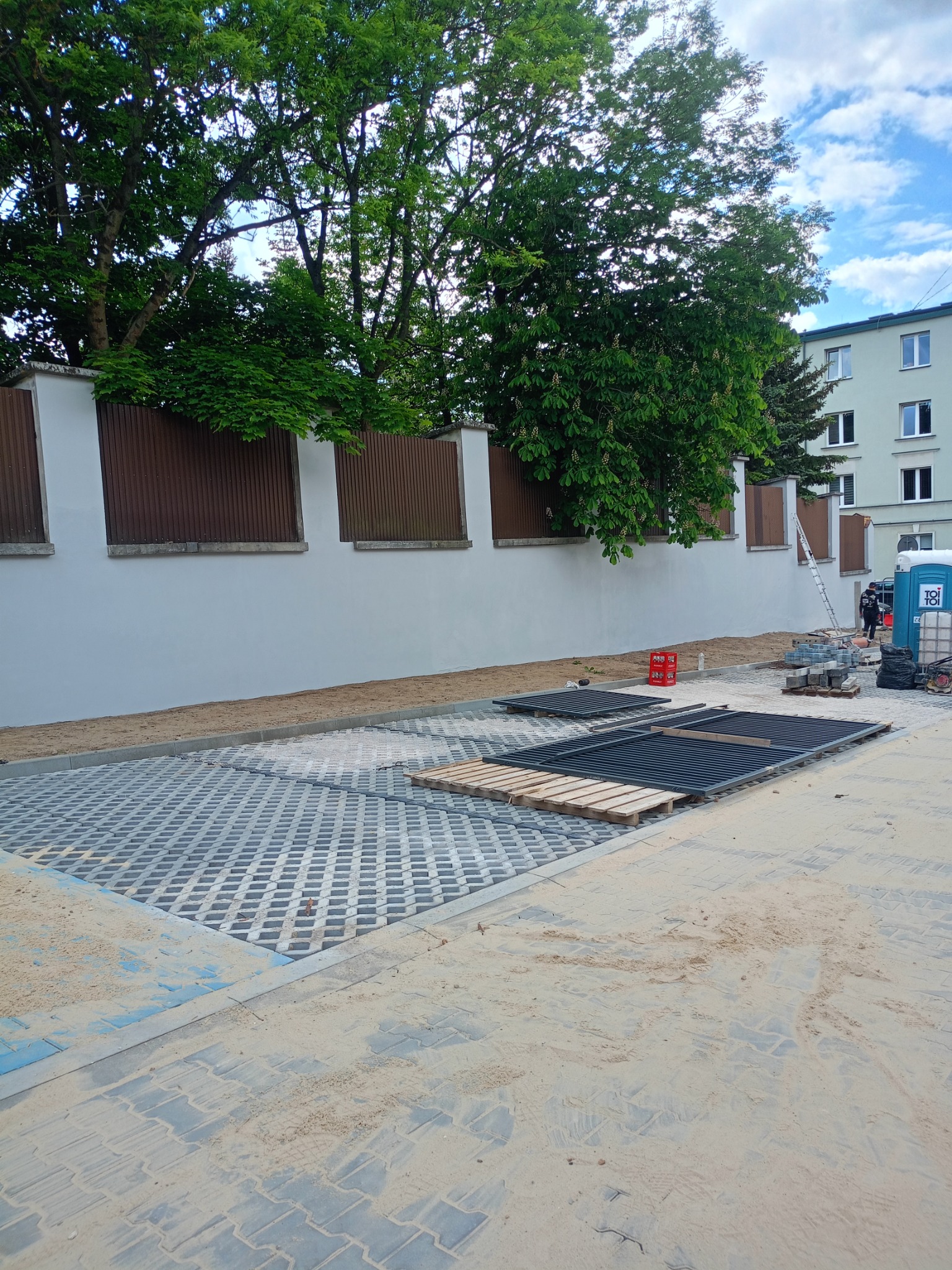 Układanie ażurowej kostki brukowej na posesji. Widoczne przygotowane elementy, palety i piasek. W tle ogrodzenie i budynek mieszkalny.