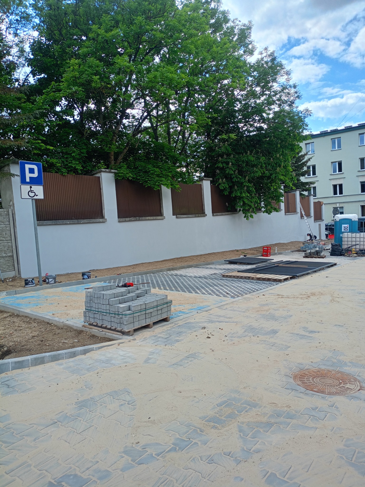 Układanie kostki brukowej na parkingu dla niepełnosprawnych. Widoczne palety z kostką, narzędzia i fragmenty ułożonej nawierzchni. Całość otoczona zielenią i budynkami.