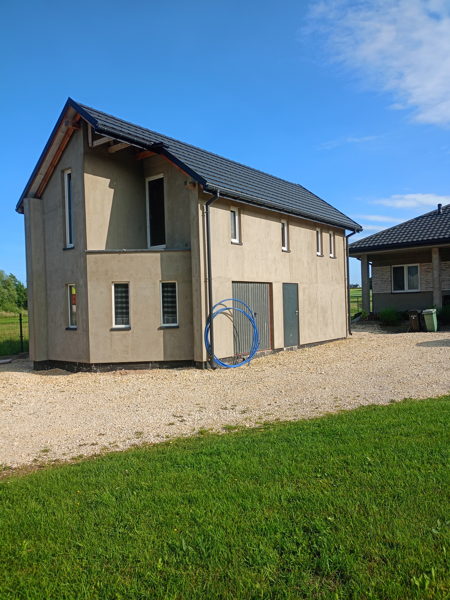 Dom z nałożoną szarą gładzią, widoczne okna z roletami. Obok leżą niebieskie rury. Na podwórku żwir i trawa. Błękitne niebo w słoneczny dzień.