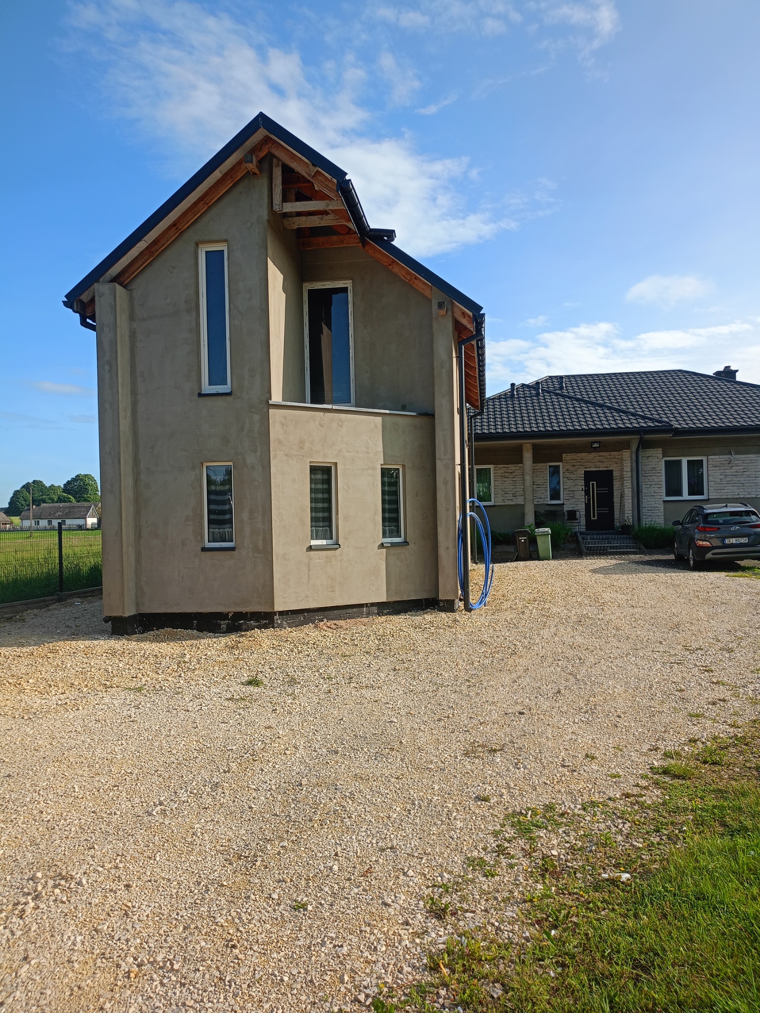 Surowa elewacja domu w trakcie budowy, z widocznymi oknami. W tle drugi budynek i niebo z chmurami. Na pierwszym planie żwirowe podłoże.