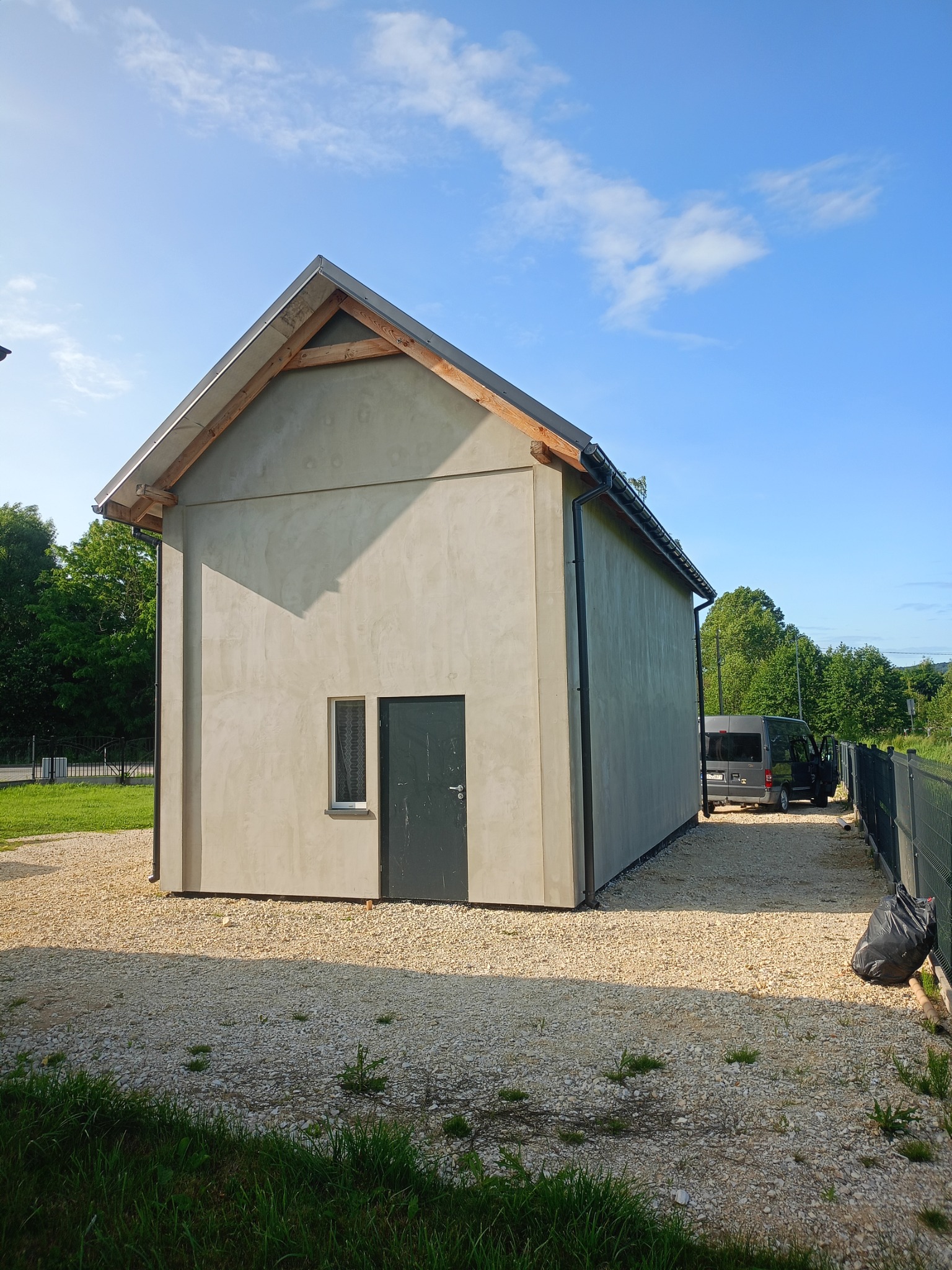 Budynek po nałożeniu szarej, gładkiej warstwy zewnętrznej, z widoczną stolarką okienną i drzwiową oraz fragmentem dachu. Obok zaparkowany bus.