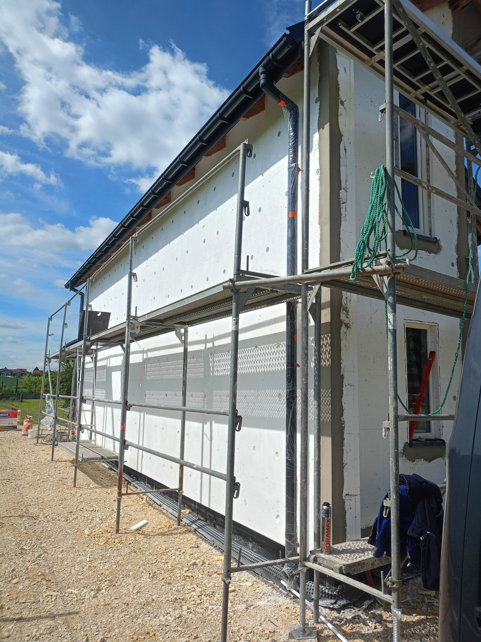 Dom w trakcie ocieplania, widoczne rusztowania, styropian z otworami, szara zaprawa, błękitne niebo z chmurami. Prace elewacyjne w toku, narzędzia na rusztowaniu.