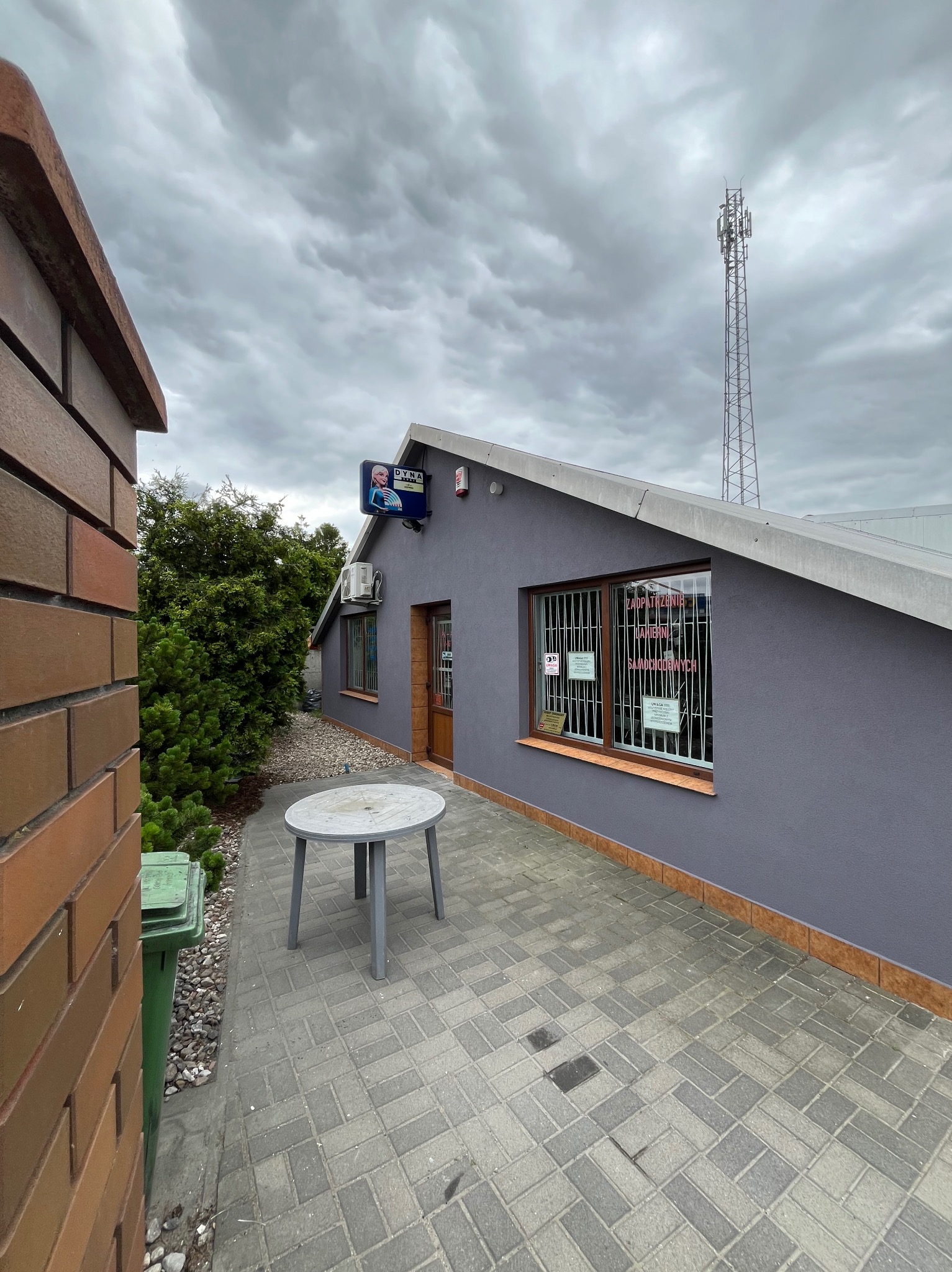 Szary budynek z oknem w kraty i reklamą, widziany z zewnątrz, z szarym stołem na pierwszym planie i pochmurnym niebem w tle. Widoczna wieża telekomunikacyjna.
