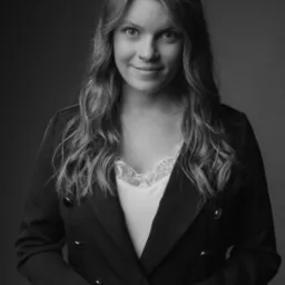 Portret kobiety w czarno-białej tonacji, ubranej w ciemną marynarkę i jasną bluzkę z koronką, z delikatnym uśmiechem i pierścionkiem na palcu.