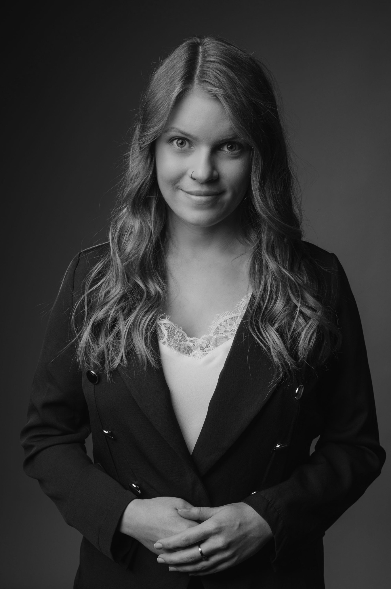Portret kobiety w czarno-białej tonacji, ubranej w ciemną marynarkę i jasną bluzkę z koronką, z delikatnym uśmiechem i pierścionkiem na palcu.