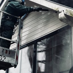 Zielony Tulipan - Pracownik w kombinezonie i masce ochronnej, stojący na podnośniku, czyści siding elewacji za pomocą myjki ciśnieniowej, widoczne zacieki i brud na elewacji.