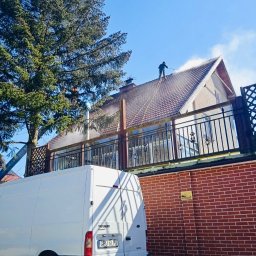 Zielony Tulipan - Pracownik w kombinezonie roboczym czyści dach domu jednorodzinnego za pomocą myjki ciśnieniowej, widoczny biały samochód dostawczy na tle ceglanego muru i drzewa.