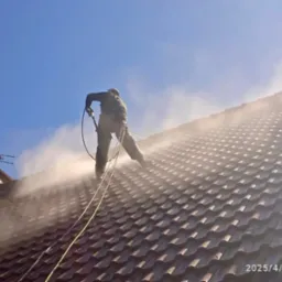 Pracownik w uprzęży myje dach z brązowej dachówki za pomocą myjki ciśnieniowej, widoczna antena telewizyjna na kominie