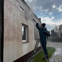 Zielony Tulipan - Pracownik w kombinezonie ochronnym na drabinie czyści elewację budynku we Władysławowie za pomocą myjki ciśnieniowej. Widoczne różnice w kolorze elewacji przed i po czyszczeniu.