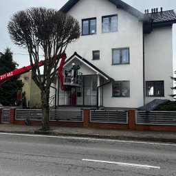 Zielony Tulipan - Mycie elewacji domu jednorodzinnego z podnośnika koszowego. Widoczna para wodna i czyszczona powierzchnia. Na pierwszym planie drzewo.