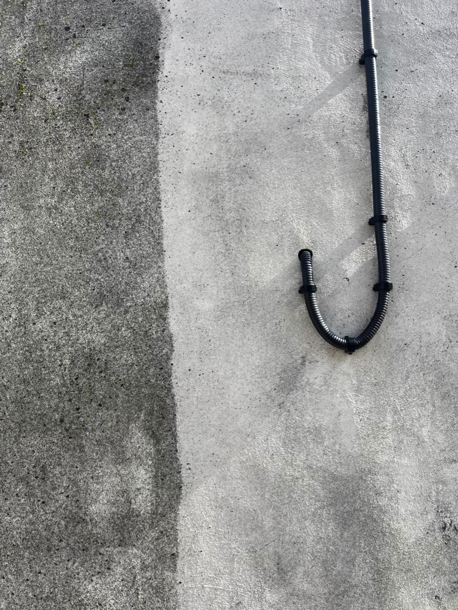 Fragment szarej elewacji z widoczną różnicą w czystości po zabiegu czyszczenia; pionowa linia oddziela brudną i czystą powierzchnię, po prawej stronie czarna rurka instalacyjna przymocowana do ściany.