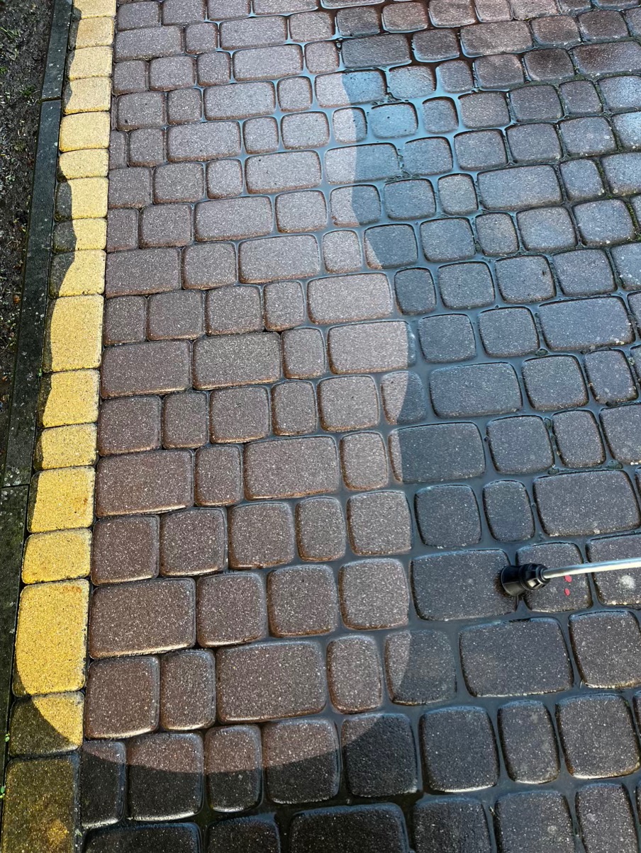 Częściowo wyczyszczona kostka brukowa z widoczną różnicą w kolorze, po lewej stronie żółta obwódka, woda na powierzchni kostki, lanca myjąca w prawym dolnym rogu.