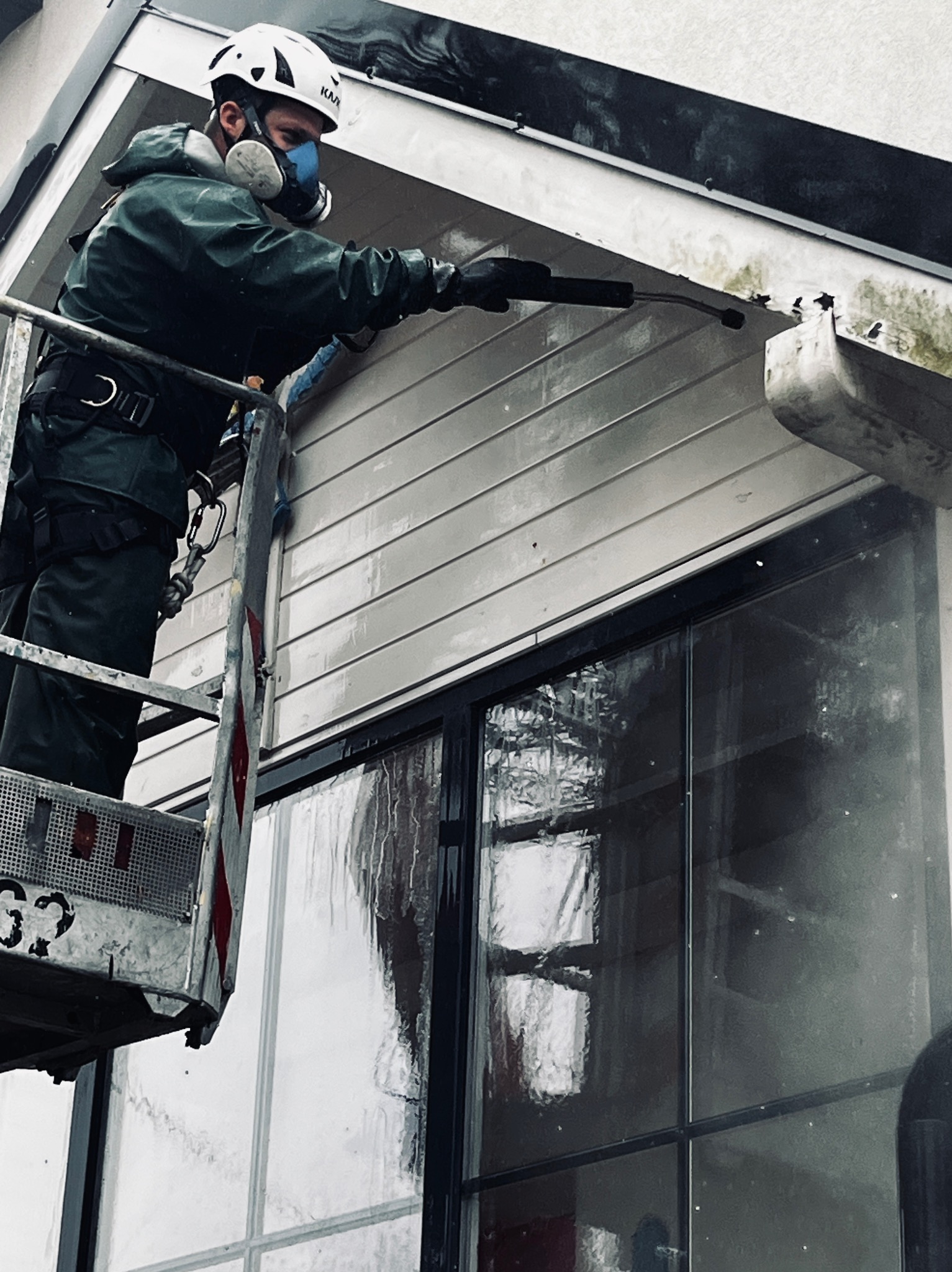 Pracownik w kombinezonie i masce ochronnej, stojący na podnośniku, czyści siding elewacji za pomocą myjki ciśnieniowej, widoczne zacieki i brud na elewacji.