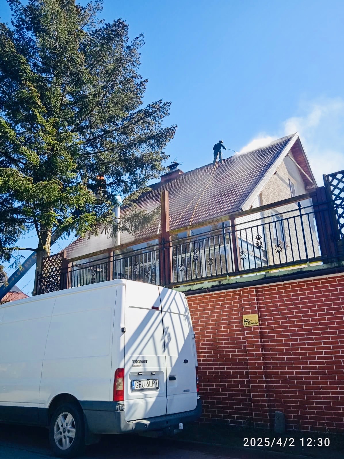 Pracownik w kombinezonie roboczym czyści dach domu jednorodzinnego za pomocą myjki ciśnieniowej, widoczny biały samochód dostawczy na tle ceglanego muru i drzewa.