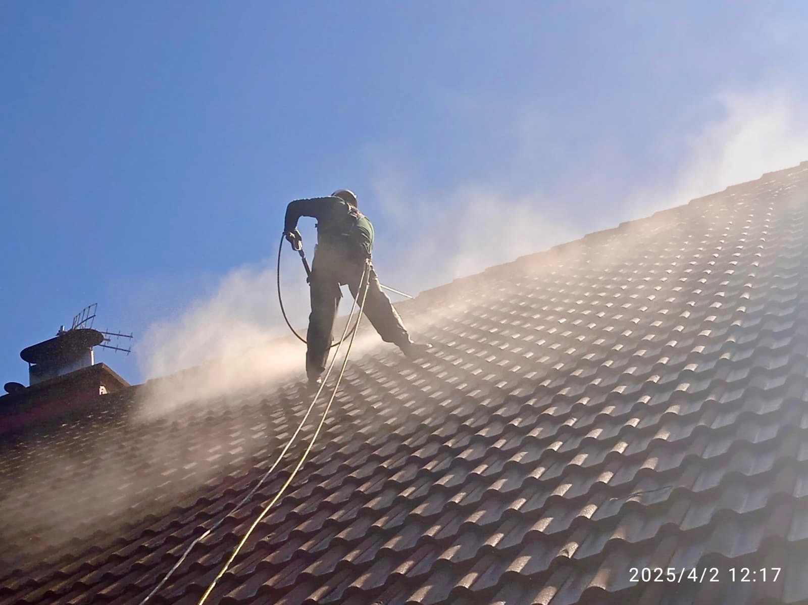 Pracownik w uprzęży myje dach z brązowej dachówki za pomocą myjki ciśnieniowej, widoczna antena telewizyjna na kominie
