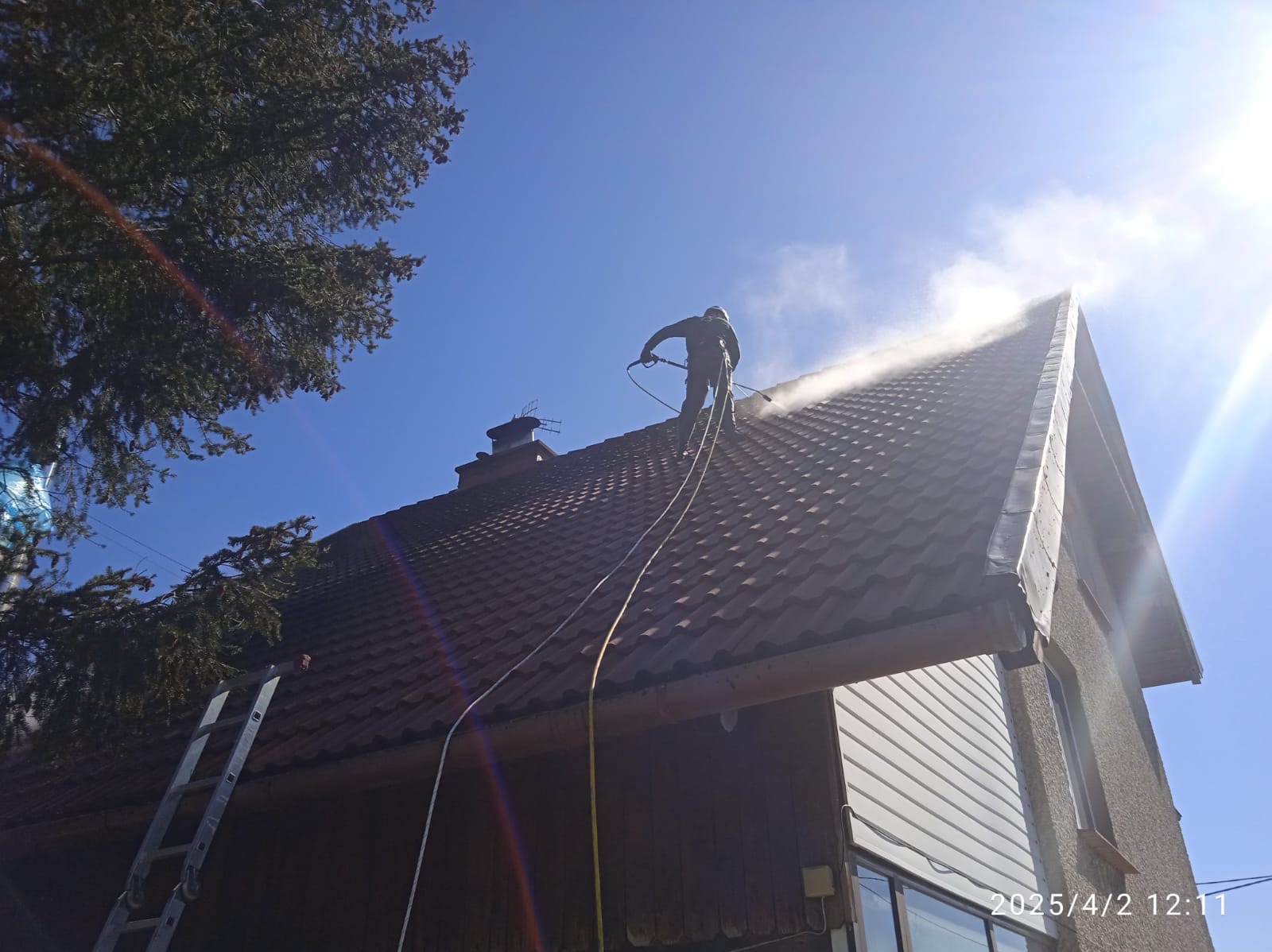 Pracownik w kombinezonie spryskuje dach z brązowej dachówki strumieniem wody pod ciśnieniem, widoczny komin i drabina oparta o budynek, błękitne niebo w tle.