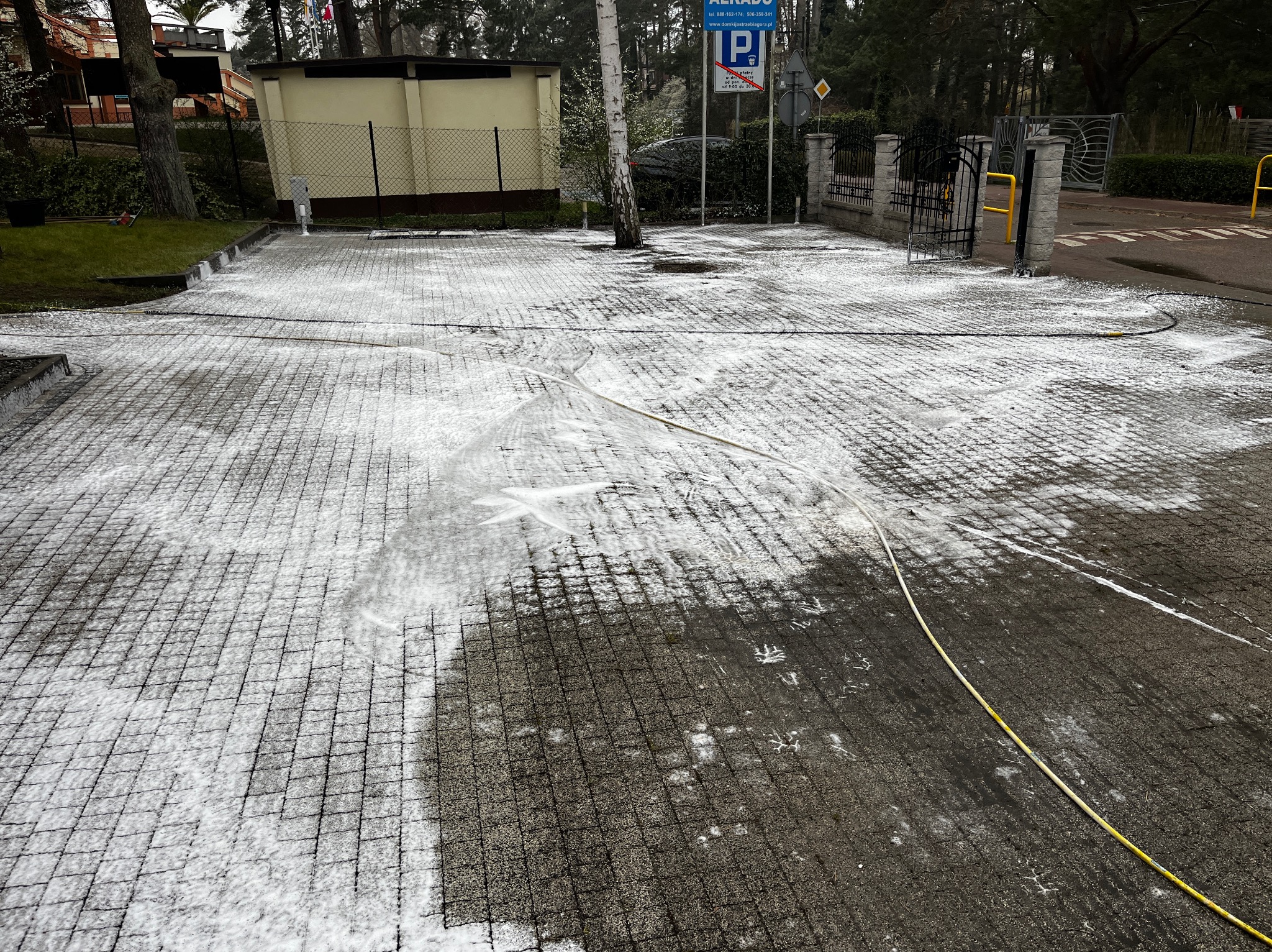 Częściowo oczyszczona kostka brukowa z resztkami piany i śladami po czyszczeniu, widoczny wąż ciśnieniowy, na tle budynek i drzewa.
