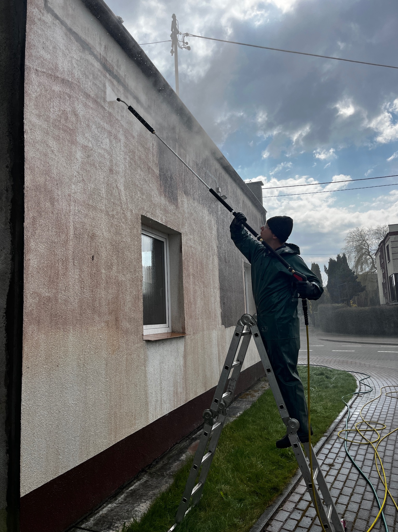 Pracownik w kombinezonie ochronnym na drabinie czyści elewację budynku we Władysławowie za pomocą myjki ciśnieniowej. Widoczne różnice w kolorze elewacji przed i po czyszczeniu.