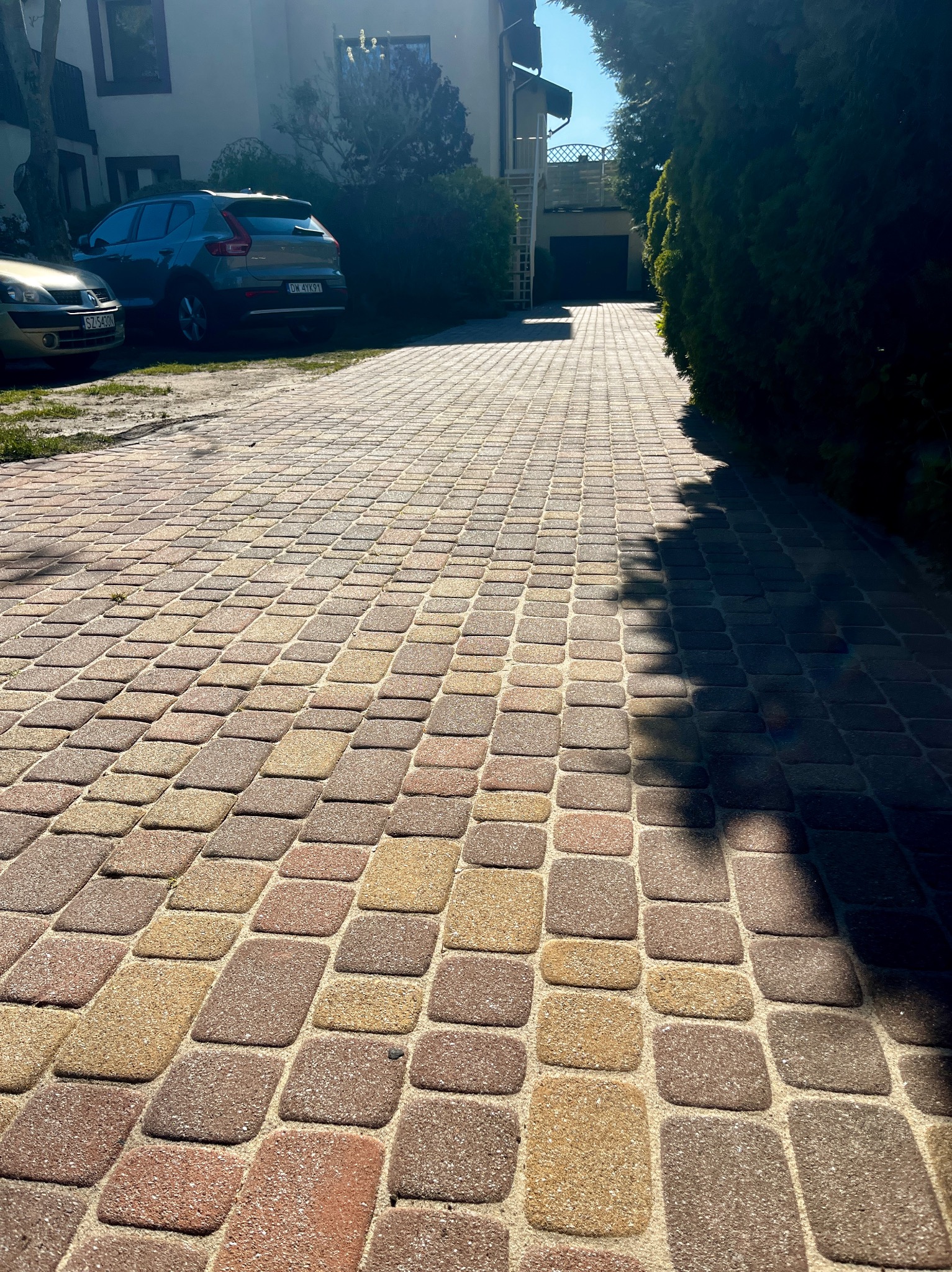 Kolorowa kostka brukowa na podjeździe domu w słoneczny dzień. Widoczne dwa auta i zieleń. Cień drzewa pada na część powierzchni. Regularny wzór kostki.