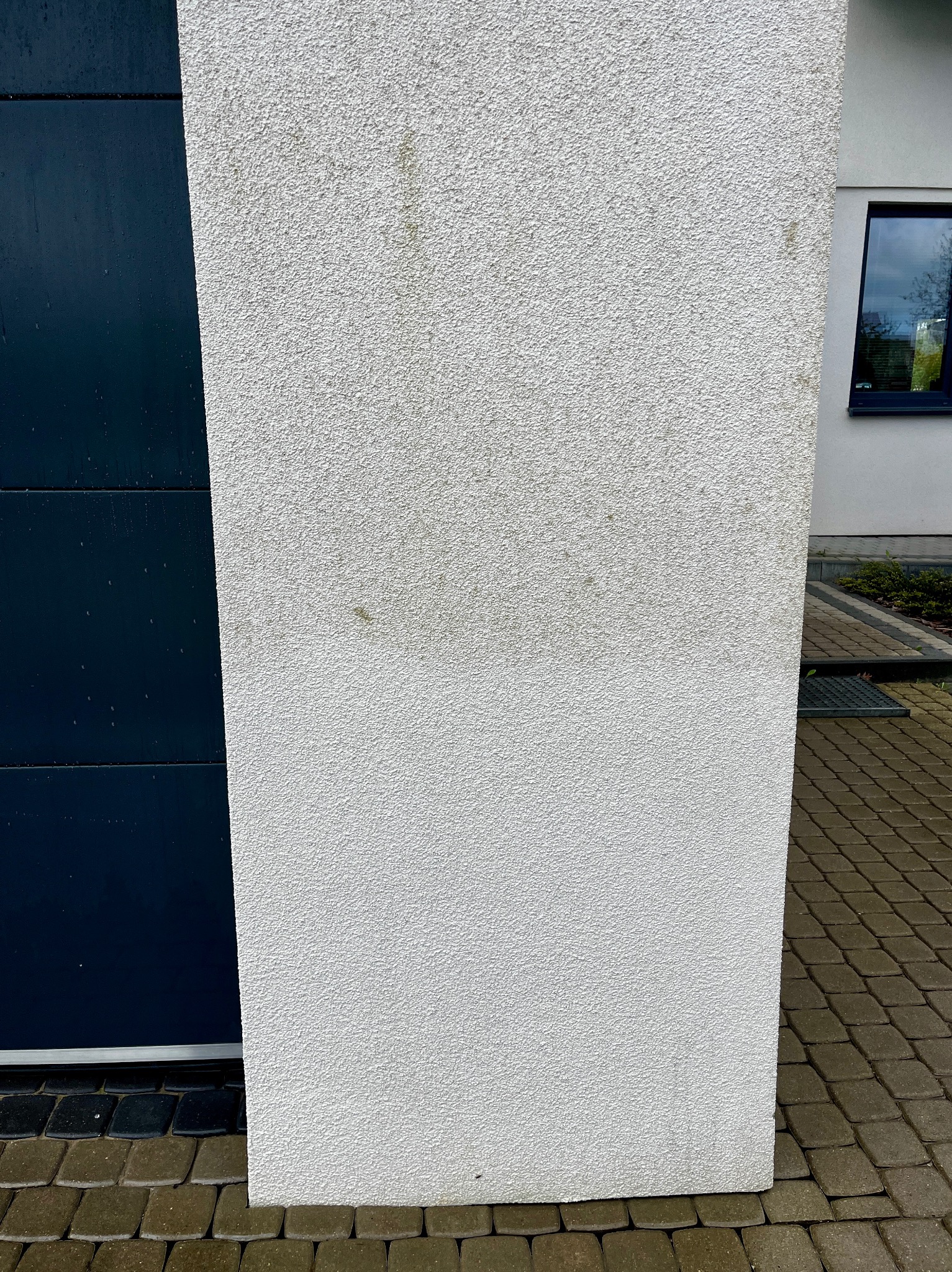 Fragment elewacji domu z widoczną różnicą w czystości, gdzie dolna część jest wyraźnie bielsza od górnej, z plamami. Obok fragment bramy garażowej i kostki brukowej.