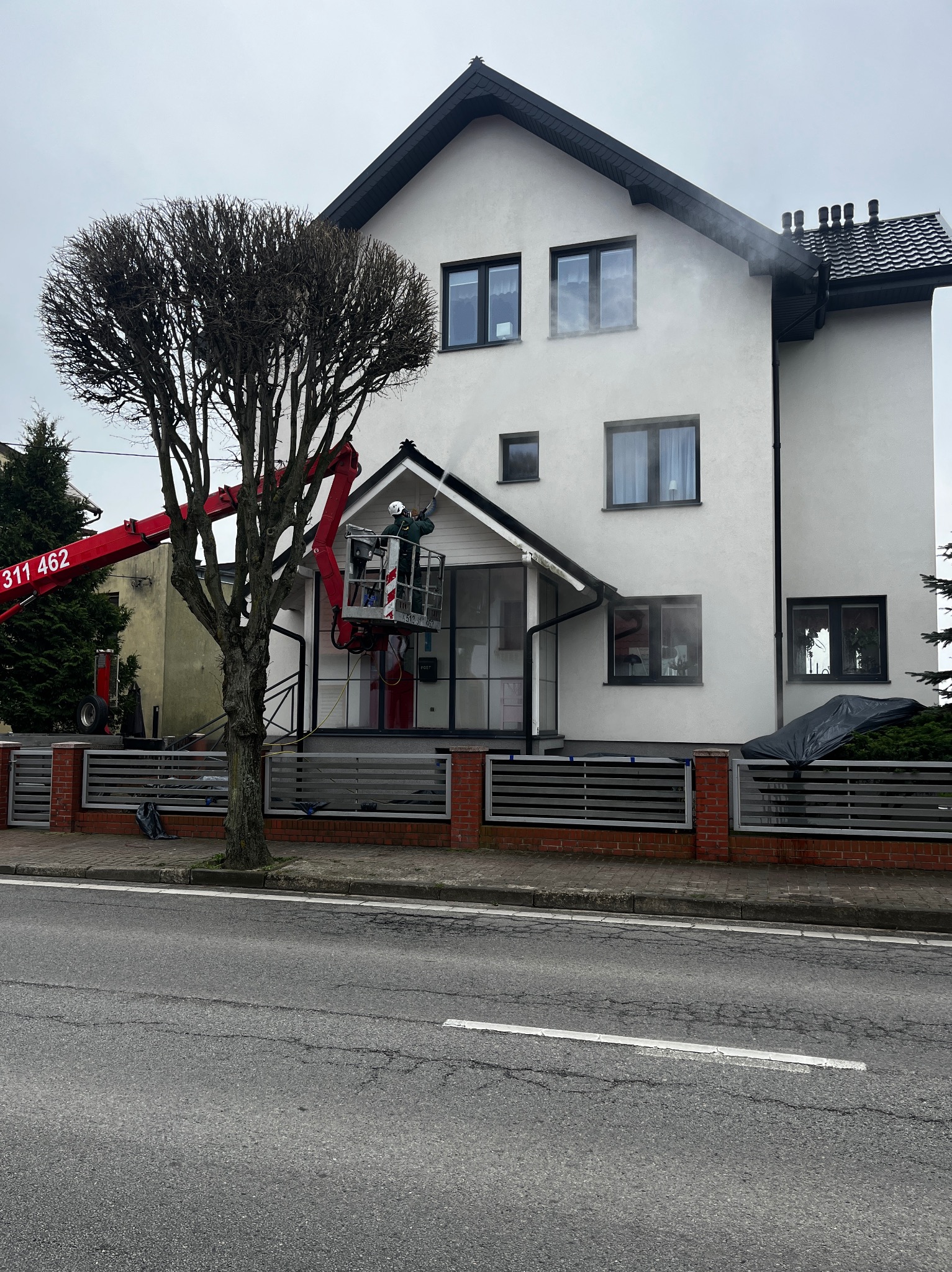 Mycie elewacji domu jednorodzinnego z podnośnika koszowego. Widoczna para wodna i czyszczona powierzchnia. Na pierwszym planie drzewo.