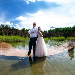Studio filmowe Heniek Lucyna Jaszczyszyn - Para w strojach ślubnych stoi na drewnianym pomoście zanurzonym w wodzie, tło stanowi skalisty brzeg porośnięty zielenią i błękitne niebo.
