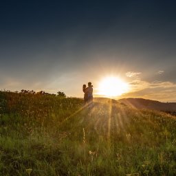 Studio filmowe Heniek Lucyna Jaszczyszyn - Para zakochanych obejmująca się na szczycie zielonego wzgórza podczas zachodu słońca, z promieniami słonecznymi przebijającymi się przez trawę.