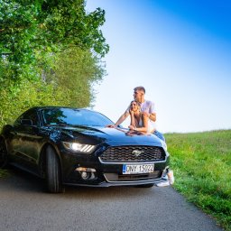 Studio filmowe Heniek Lucyna Jaszczyszyn - Para oparta o czarnego Forda Mustanga z polskimi tablicami rejestracyjnymi ONY 15022 na tle zieleni i błękitnego nieba, ujęcie z dołu.