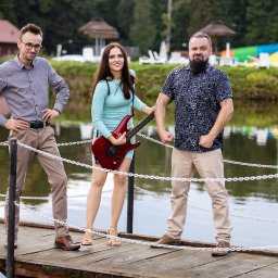 Studio filmowe Heniek Lucyna Jaszczyszyn - Zespół muzyczny pozuje na drewnianym pomoście nad jeziorem, w tle widać budynki rekreacyjne i park wodny. Kobieta w środku trzyma czerwoną gitarę elektryczną.