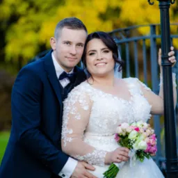 Para młoda w Prudniku pozuje do zdjęcia na tle metalowego mostku i jesiennych drzew, panna młoda w koronkowej sukni z bukietem, pan młody w granatowym garniturze z muszką.