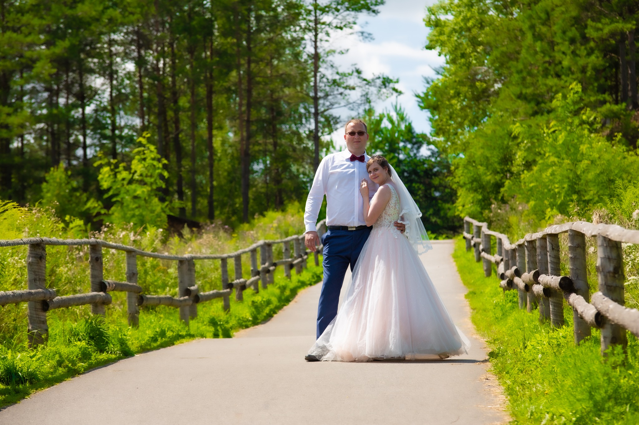 Para młoda pozuje na leśnej ścieżce z drewnianą barierką; pan młody w białej koszuli, granatowych spodniach i bordowej muszce, panna młoda w białej sukni ślubnej z koronkową górą i welonem.