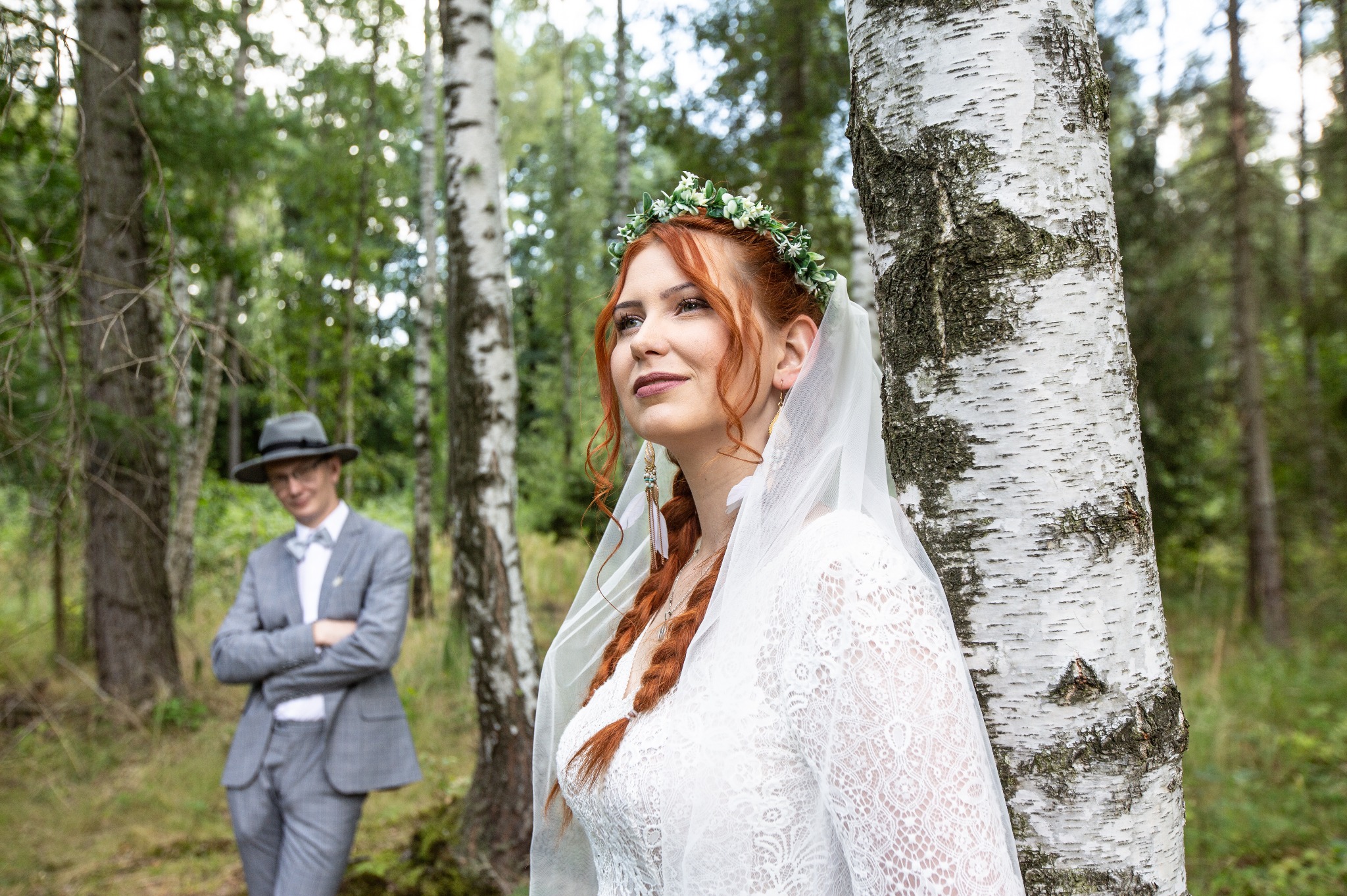 Panna młoda z rudymi warkoczami, wiankiem i welonem, oparta o brzozę w lesie, obok stoi pan młody w szarym garniturze i kapeluszu.